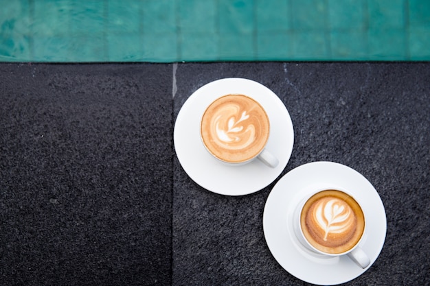 Twee witte kopjes lekkere cappucino met art latte aan de rand van het zwembad. vakantie tijd concept.