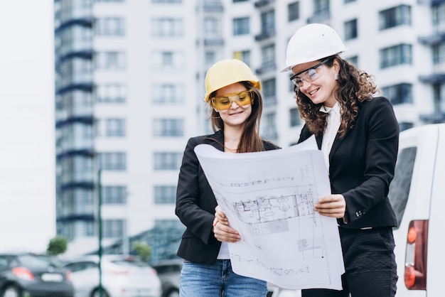 Twee zakelijke vrouwen in beschermende helmen en veiligheidsbril kijken naar bouwschema&#39;s, architectonisch concept