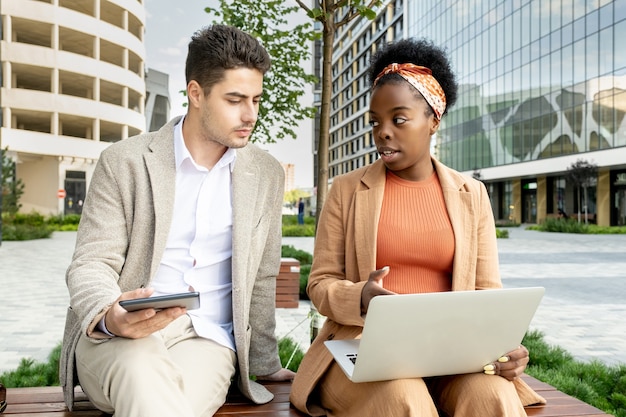 Twee zakenpartners zittend op een bankje met laptop en online werken in team in de stad