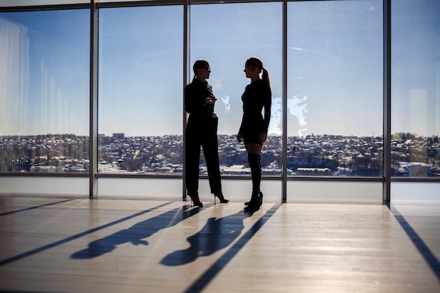 Twee zakenvrouwen genieten van het uitzicht op de stad en praten terwijl ze bij het grote raam op kantoor staan