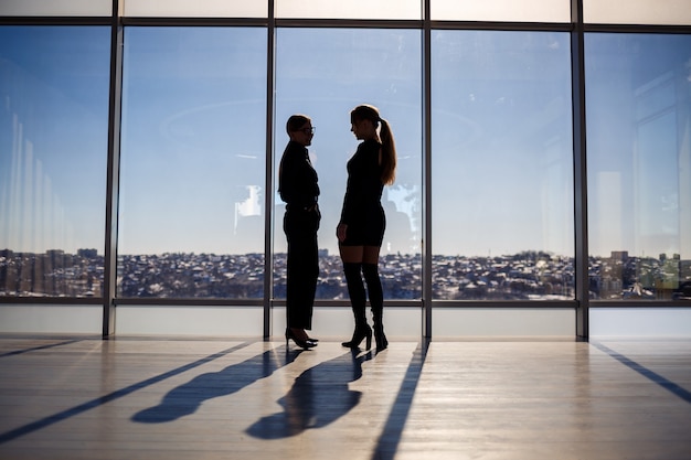 Twee zakenvrouwen genieten van het uitzicht op de stad en praten terwijl ze bij het grote raam op kantoor staan