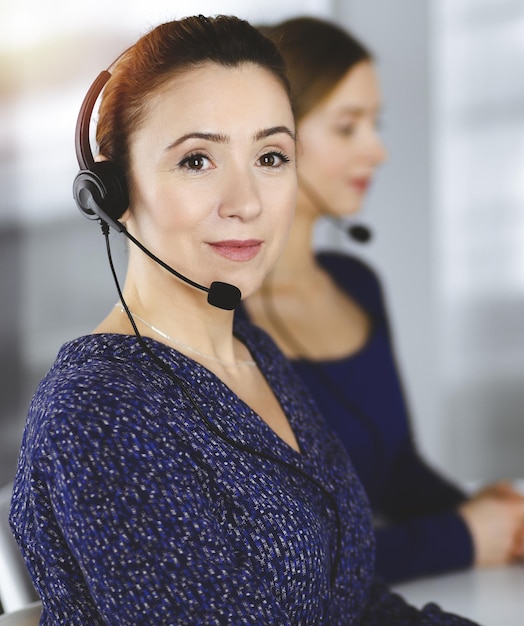 Twee zakenvrouwen voeren gesprekken met de klanten via headsets, zittend aan het bureau in een zonnig modern kantoor. Diverse mensen groeperen in een callcenter. Telemarketing en klantenservice