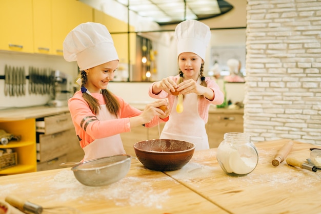 Twee zusjes koken in caps kneedt eieren in een kom, koekjes voorbereiding op de keuken. Kinderen koken gebak, kinderen chef-koks maakt deeg, kind voorbereiding van cake