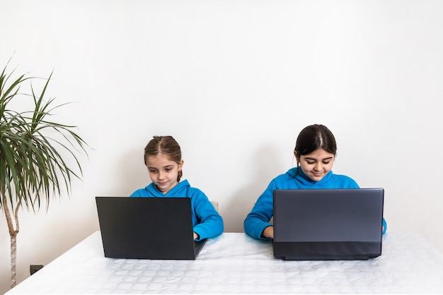 Twee zussen, een blonde en een brunette thuis in schooluniform met blauwe sweater die thuis huiswerk aan het studeren is met laptop