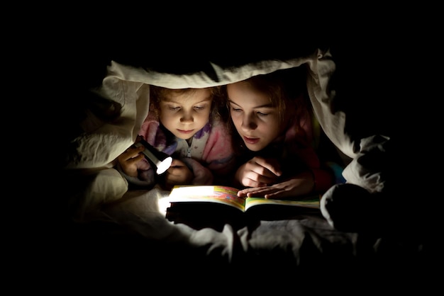 Twee zussen lezen 's nachts een boek onder de dekens met een zaklamp