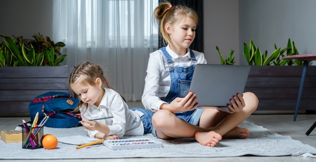 Twee zussen liggend op de vloer in de woonkamer met behulp van laptop, spelen en tekenen