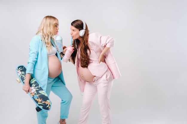 Twee zwangere meisjes in turquoise en roze pakken met glazen sap, een skate en koptelefoon staan op een grijze achtergrond