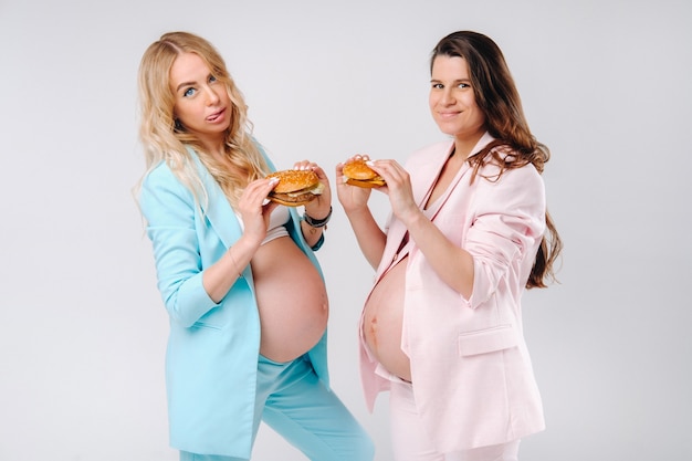 Twee zwangere vrouwen in pakken met hamburgers in hun handen op een grijze achtergrond