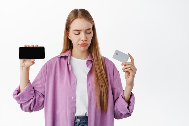 Twijfelachtige jonge vrouw die een horizontaal smartphonescherm toont en sceptisch kijkt naar de bankcreditcard die denkt een keuze te maken die over een witte achtergrond staat