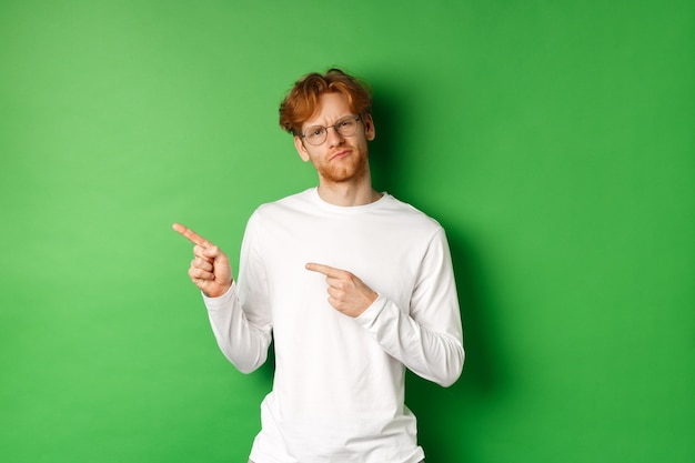 Twijfelachtige roodharige man met een bril die met de vingers naar links wijst, aarzelend en onzeker naar de camera kijkt, staande over een groene achtergrond.