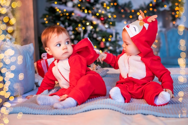 Twins toddlers in red santa's reindeer suits sit next to each other at home against the background of christmas tree