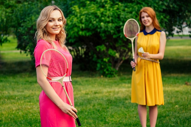 Two active cheerful girlfriends beautiful young ginger redhead irish woman in a yellow dress and european blonde female person in a pink dress playing tennis badminton racket in the summer park