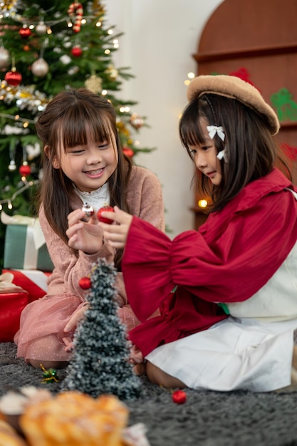 Two adorable Asian girls are enjoying playing and decorating a small Christmas tree in a living room