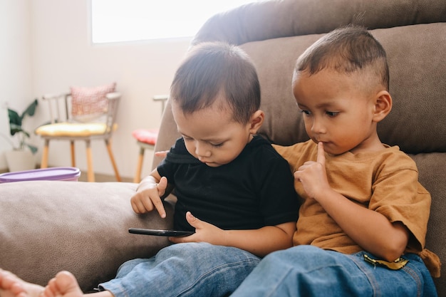Photo two adorable asian little boys sitting on sofa and playing smartphone