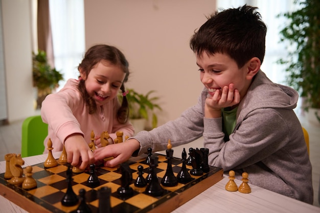 Two adorable kids, boy and girl, brother and sister having great time playing chess game together at home interior.