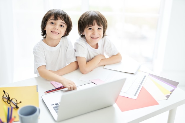 Two adorable latin boys, brothers smiling at camera while sitting together at the table and using laptop. Little kids having online lesson at home. Children, e learning concept