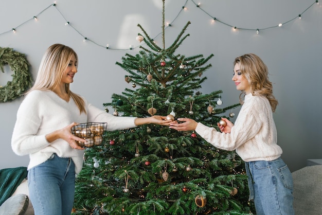 セーターとジーンズを着た 2 人の大人の女性姉妹が家でクリスマス ツリーを飾る