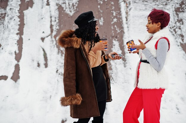 シープスキンと毛皮のコートで2人のアフリカ系アメリカ人女性が冬の日にカップで雪を背景にポーズ
