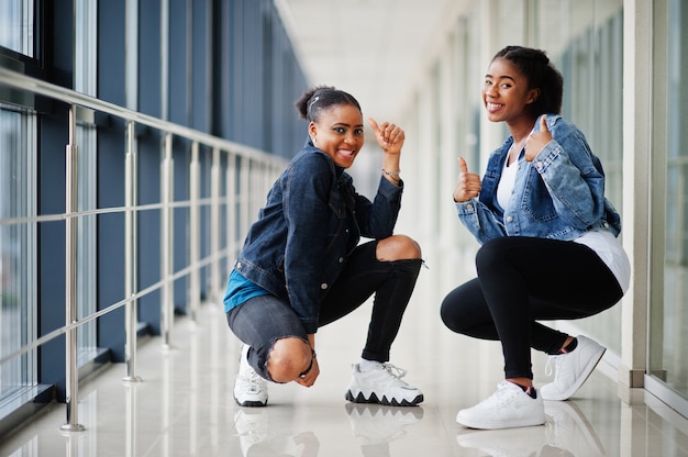 Two african woman friends in jeans jacket show thumb up indoor together.