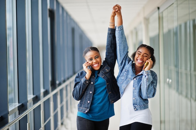 Two african woman friends in jeans jacket speak on phone and hands up indoor together.