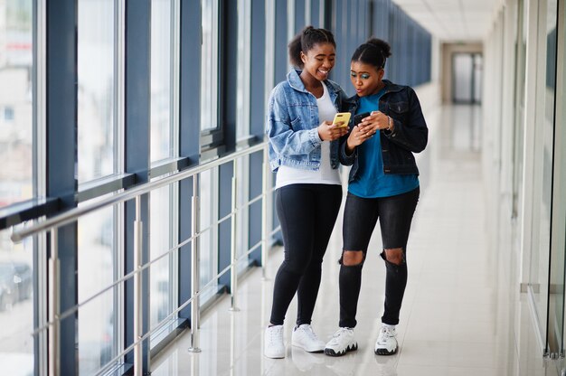 Two african woman friends in jeans jacket using mobile phones indoor together.