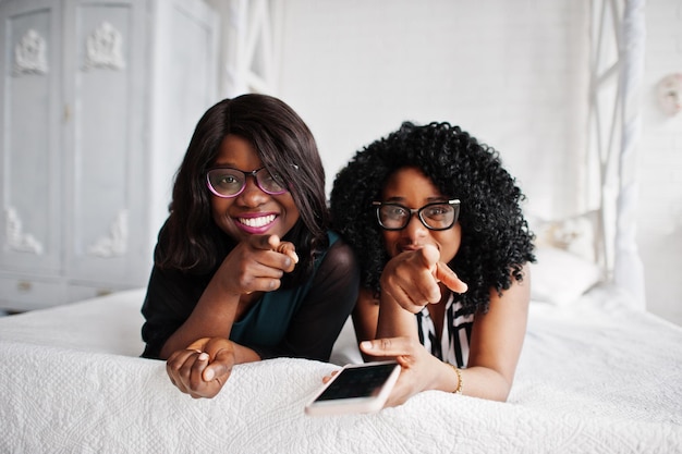 Two african woman friends wear on eyeglasses lying on the bed indoor white room and looking at mobile phone They show fingers to camera