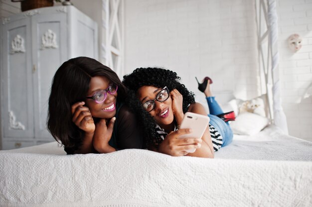 Two african woman friends wear on eyeglasses lying on the bed indoor white room and looking at mobile phone.