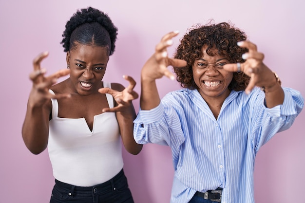 Two african women standing over pink background shouting frustrated with rage, hands trying to strangle, yelling mad