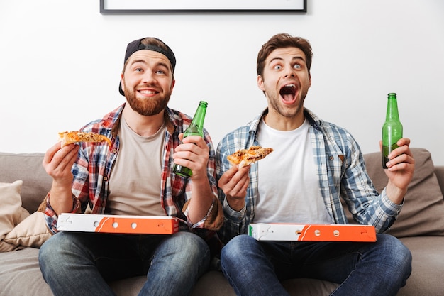 Two agitated emotional men eating pizza and drinking beer with, while supporting football team at home