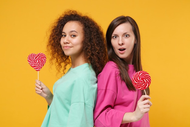 Photo two amazed women friends european and african american girls in pink green clothes posing isolated on yellow background. people lifestyle concept. mock up copy space. holding round heart lollipops.