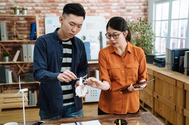 Two architects asian japanese man and woman working on\
construction project in office. young male and female coworkers\
standing at wooden table in eco friendly workplace discussing on\
model windmill