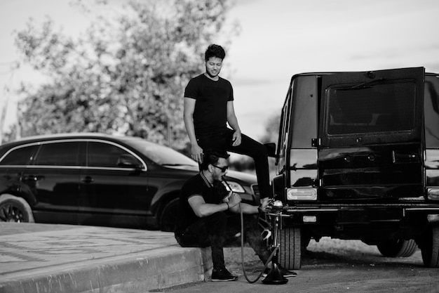 Two asian brothers man wear on all black posed near suv cars and smoke hookah.