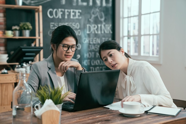 two asian businesswomen having conversation discussion working in cafe bar in morning waiting for job interview. elegant office colleague talking with new project on laptop computer in coffee shop.