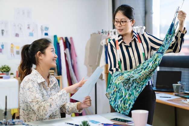 Two asian chinese female fashion designers working together at\
workplace for sewing clothes. group of woman creative teamwork\
partners brainstorming over new dress designs. lady showing\
products