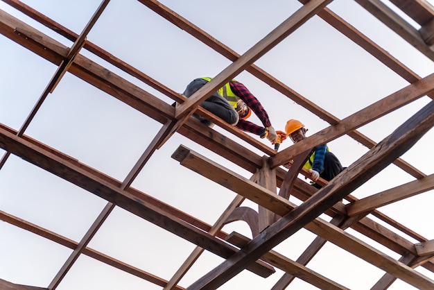 2人のアジアの建設労働者が新しい屋根、屋根ふきツール、木製の屋根構造の新しい屋根に使用される電気ドリル、チームワーク建設コンセプトをインストールします。