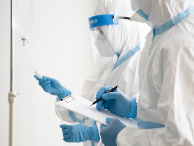 Two asian doctor wear PPE suit with N95 mask and face shield, hold patient chart and treat coronavirus infected patient in negative pressure room.