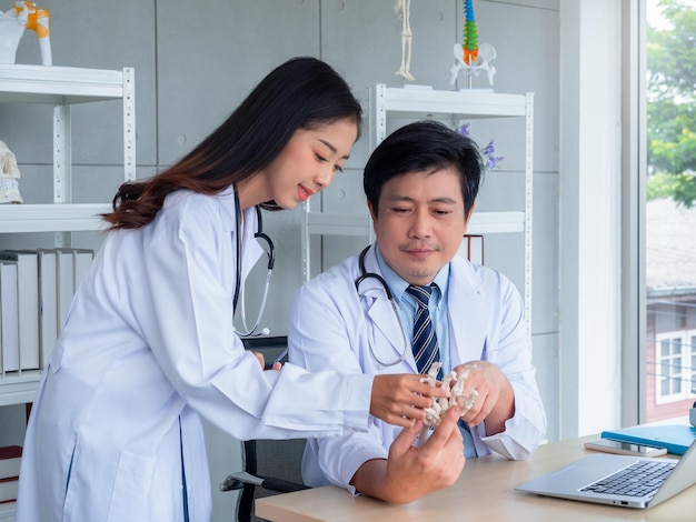 Two Asian doctors in white uniforms talking and working together in medical office The adult male doctor provides knowledge and counseling to female doctor about hand or wrist bone skeleton on desk