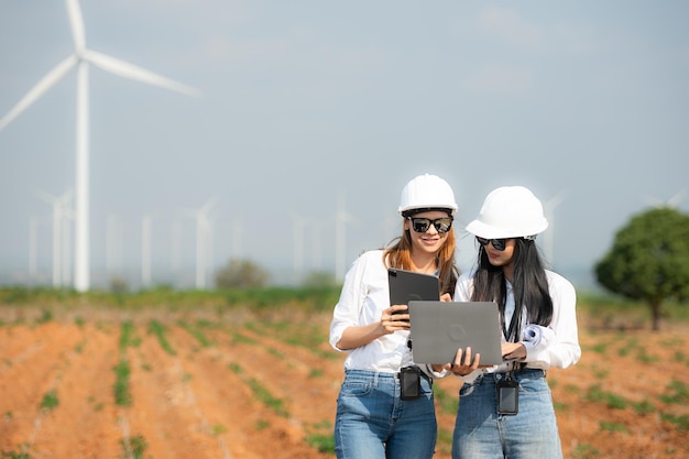 Two asian engineer women holding laptop and working in wind turbine farm with blue sky background