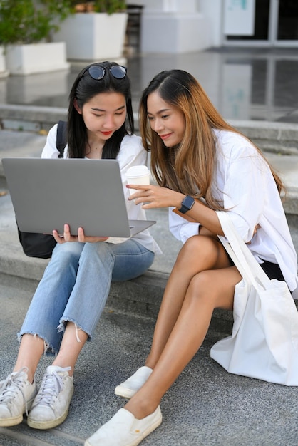 Two Asian female college students sitting on stairs and looking something at laptop screen together