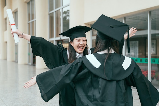 two asian girls graduates in mantles with diplomas in hands are standing near university building in hall and opening arms ready hugging each other. friendship real moments. summer graduation season.