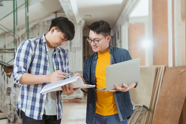 Due appaltatori maschi interni asiatici consultano ed esaminano la ristrutturazione della casa del sito con un ingegnere e designer asiatico di disegno di laptop e carta spiegano e discutono durante la costruzione del sito