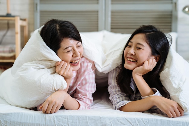 두 명의 동양인 한국의 가장 친한 친구가 침대에서 비밀을 이야기합니다. 잠옷을 입은 예쁜 젊은 여성들이 아늑한 침실에서 하얀 담요 아래에 누워 이야기를 나누고 있습니다. 함께 웃고 있는 아름다운 여성