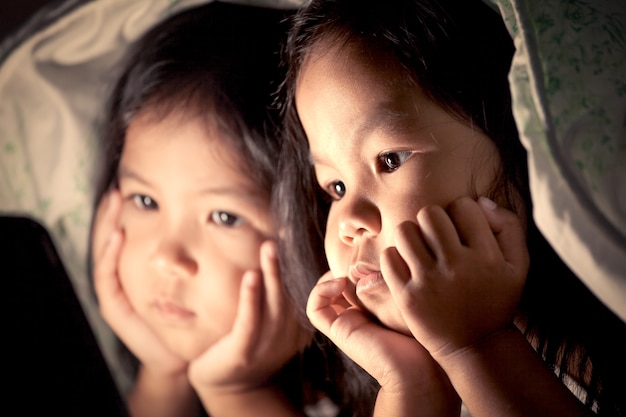 Two asian little girls using digital tablet under blanket at night in vintage color tone