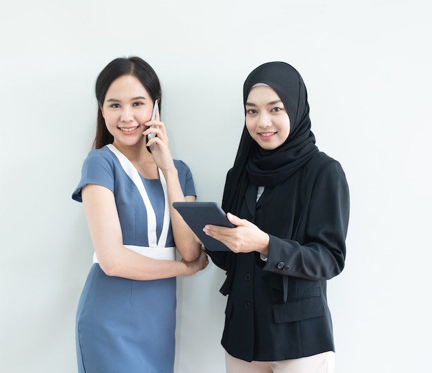 アジアのイスラム教徒の女性の2つは、スマートフォン、混合レース協力ビジネスコンセプトとモダンなスーツでタブレットと若いアジアのビジネス女性のドレスを保持します。