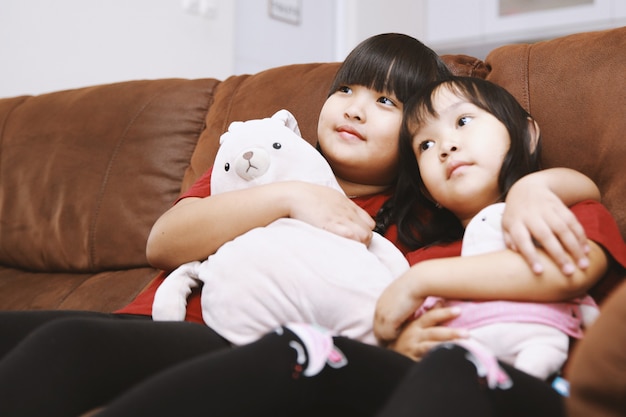 Two asian sisters sitting on couch with doll watching TV at home