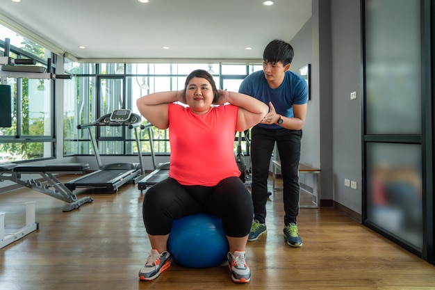 Two Asian trainer man and Overweight woman exercising with ball together in modern gym, happy and smile during workout. Fat women take care of health and want to lose weight concept.