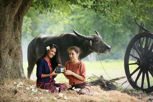 Two Asian woman wearing traditional thai costume sitting in field, while listening to vintage style radio next to buffalo