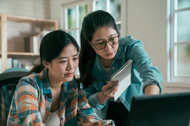 Photo two asian women colleagues working together with laptop at table in bright office. girl in glasses showing on laptop monitor with pen in hand female coworker typing keyboard. teamwork concept.