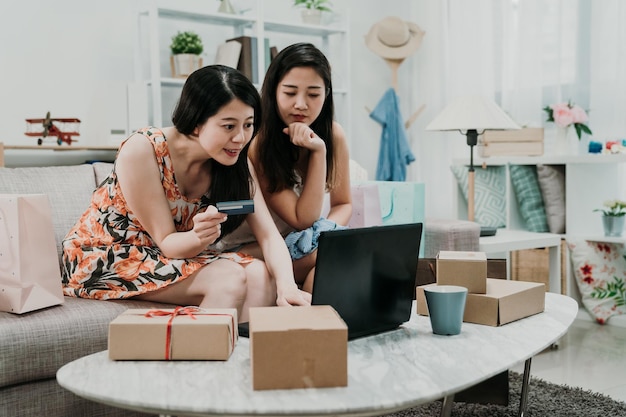 Two asian women sitting in sofa holding credit card and using laptop computer shopping online at home with products in cardboards on table. joy girl friends buying purchasing on bargain summer sale.