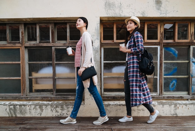 두 명의 아시아 여성 관광객이 오래된 집이 있는 기념비적인 작은 마을을 관광하고 있습니다. 나무 바닥 야외 태양 플레어 좋은 날씨에 가기 위해 커피 컵과 함께 줄을 걷는 소녀 배낭의 전체 길이.